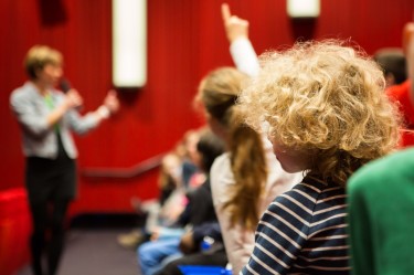 Ein roter Kinosaal. Links steht eine Person mit Mikrofon, die zum jungen Publikum spricht. Rechts sitzen mehrere Kinder in einer Reihe. Ein Kind mit langem Zopf meldet sich.